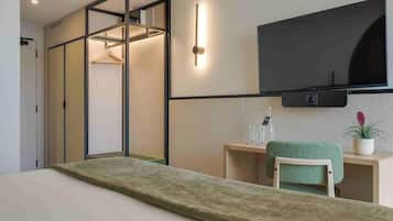 A modern hotel room with a bed, a desk, a chair, a TV mounted on the wall, and a sliding wardrobe.