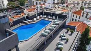 A rooftop swimming pool with lounge chairs and umbrellas, surrounded by buildings with red-tiled roofs.