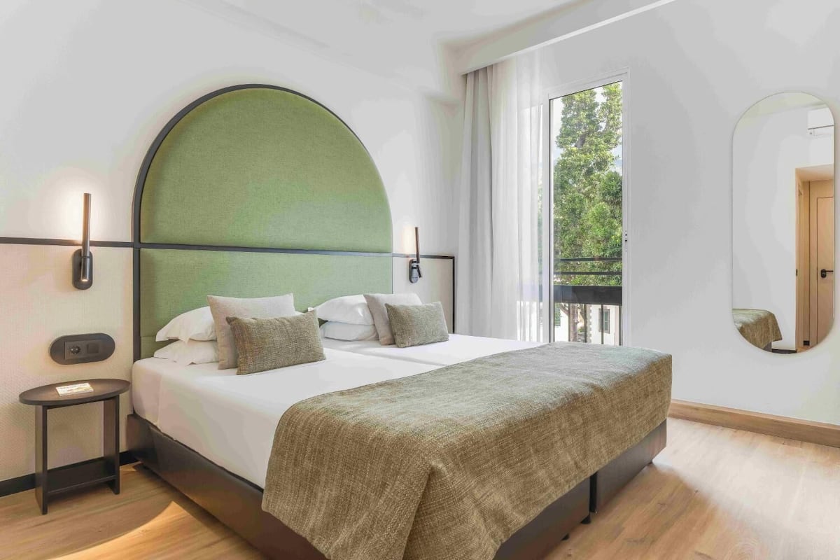 A modern bedroom with a large bed, a green headboard, a bedside table, and a large window with a view of trees.