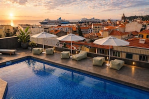Una piscina en la azotea con sillones de descanso y sombrillas, con vistas a un paisaje urbano con edificios y un atardecer.
