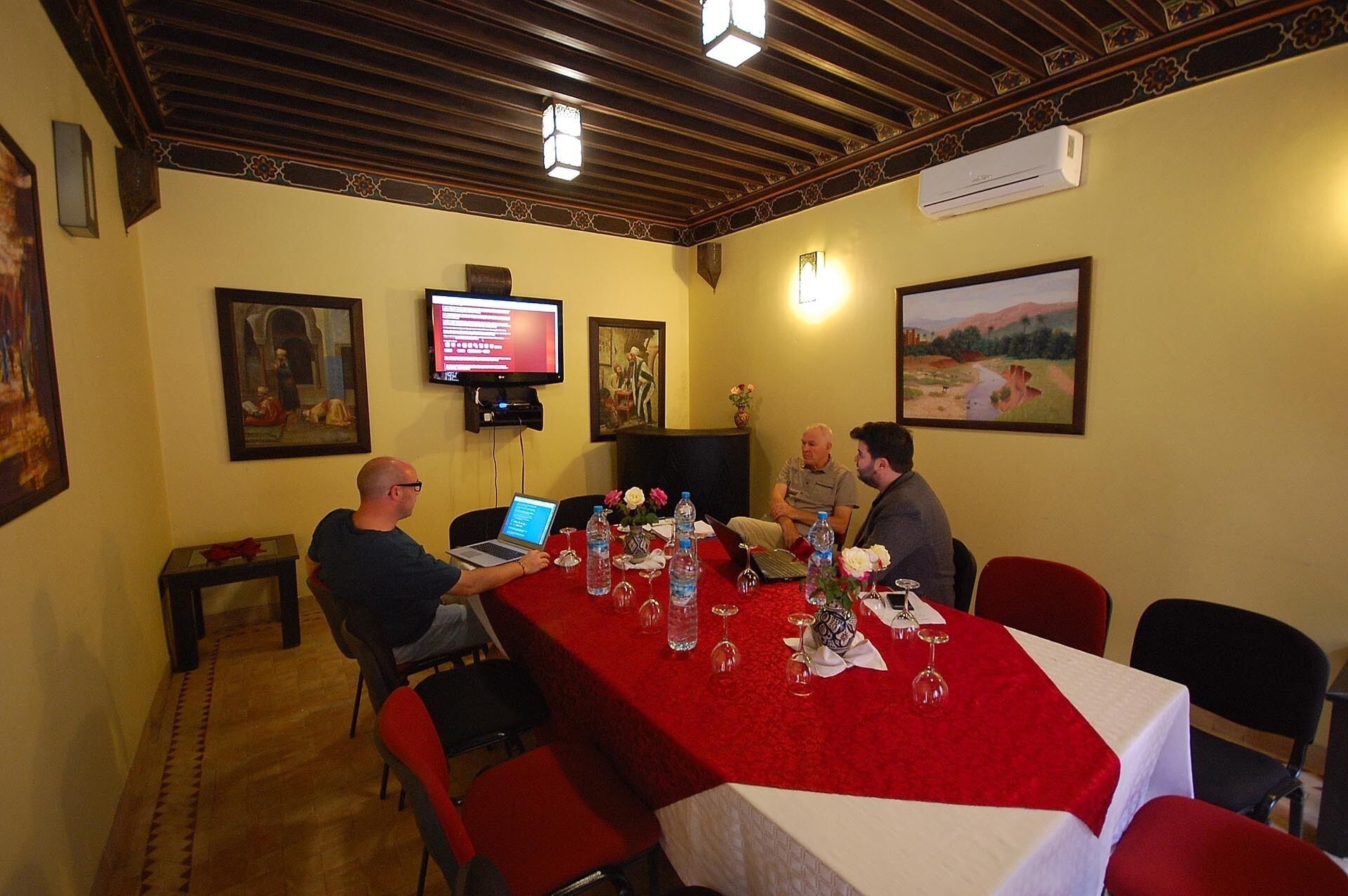 Une salle de conférence avec une longue table, des chaises rouges et un ordinateur portable posé sur la table. Des tableaux ornent les murs et un appareil de climatisation est installé au-dessus de la table.