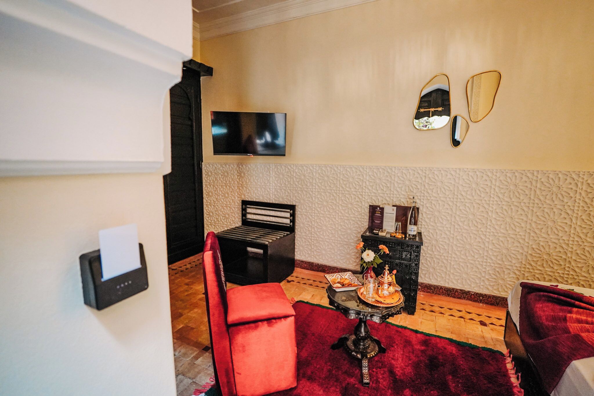 Ein Zimmer mit Flachbildfernseher, einem roten Sessel, einem kleinen Tisch mit Tablett und einem dekorativen Spiegel an der Wand.