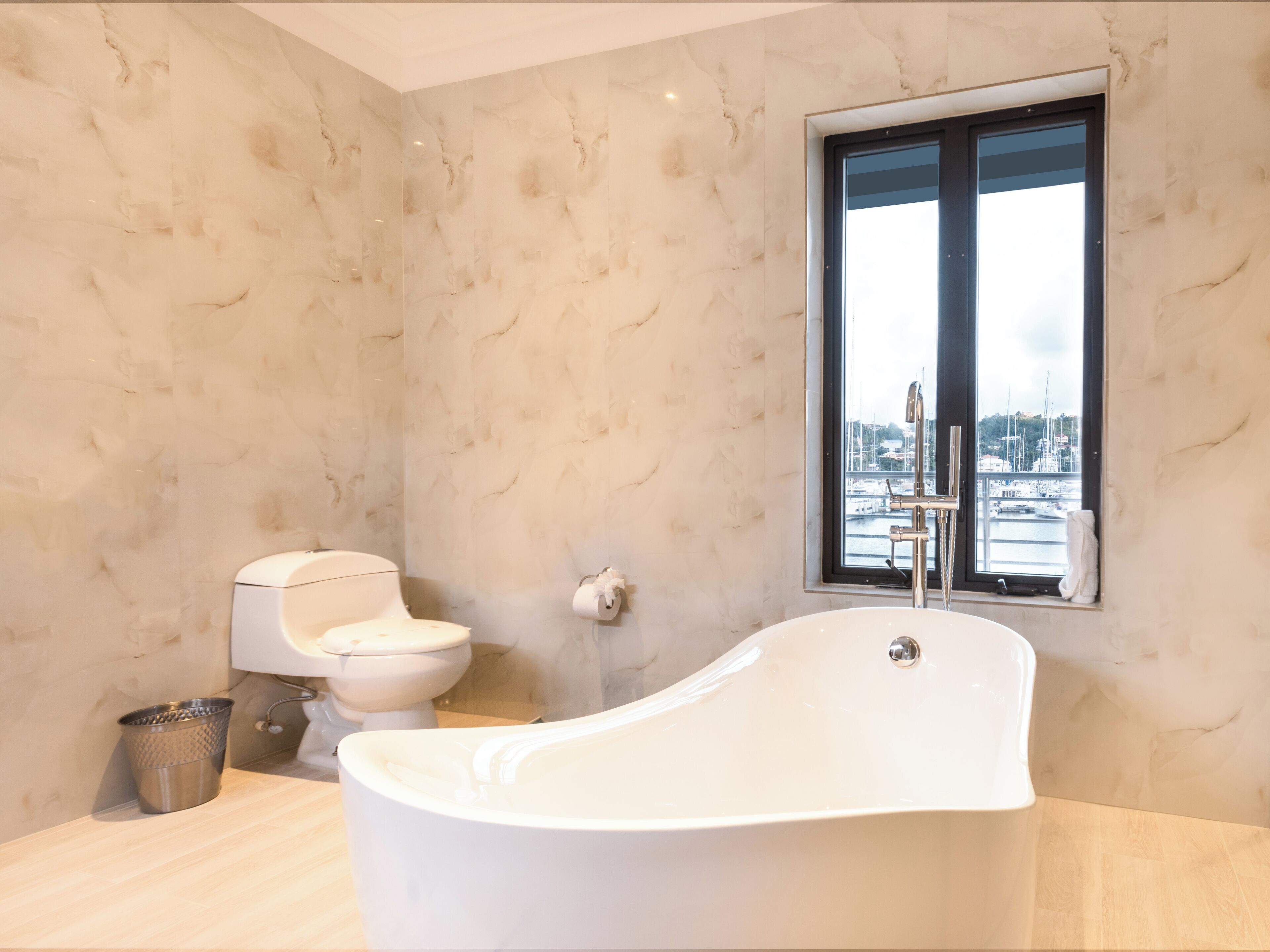 A modern bathroom with a freestanding bathtub, a toilet, and a window with a view of a marina.