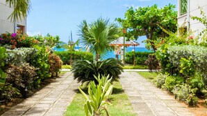 A paved pathway flanked by lush greenery and a building with a view of the sea.