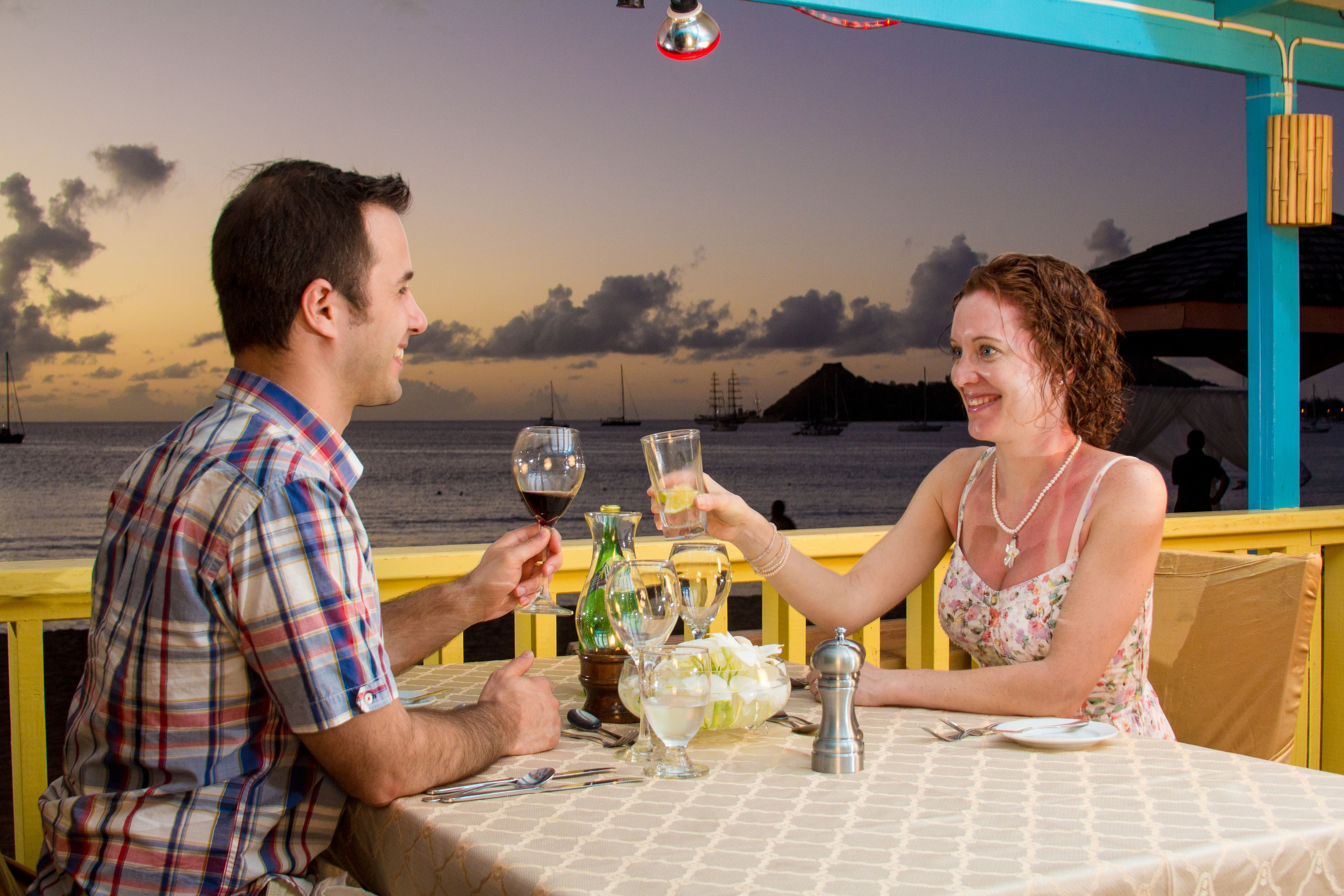 A couple toasting at a table with a view of the sea and boats in the background.