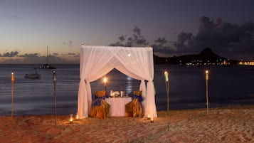 Una carpa en la playa con una mesa dispuesta para dos personas, rodeada de velas, con vistas a aguas tranquilas, barcos y una montaña en la distancia.