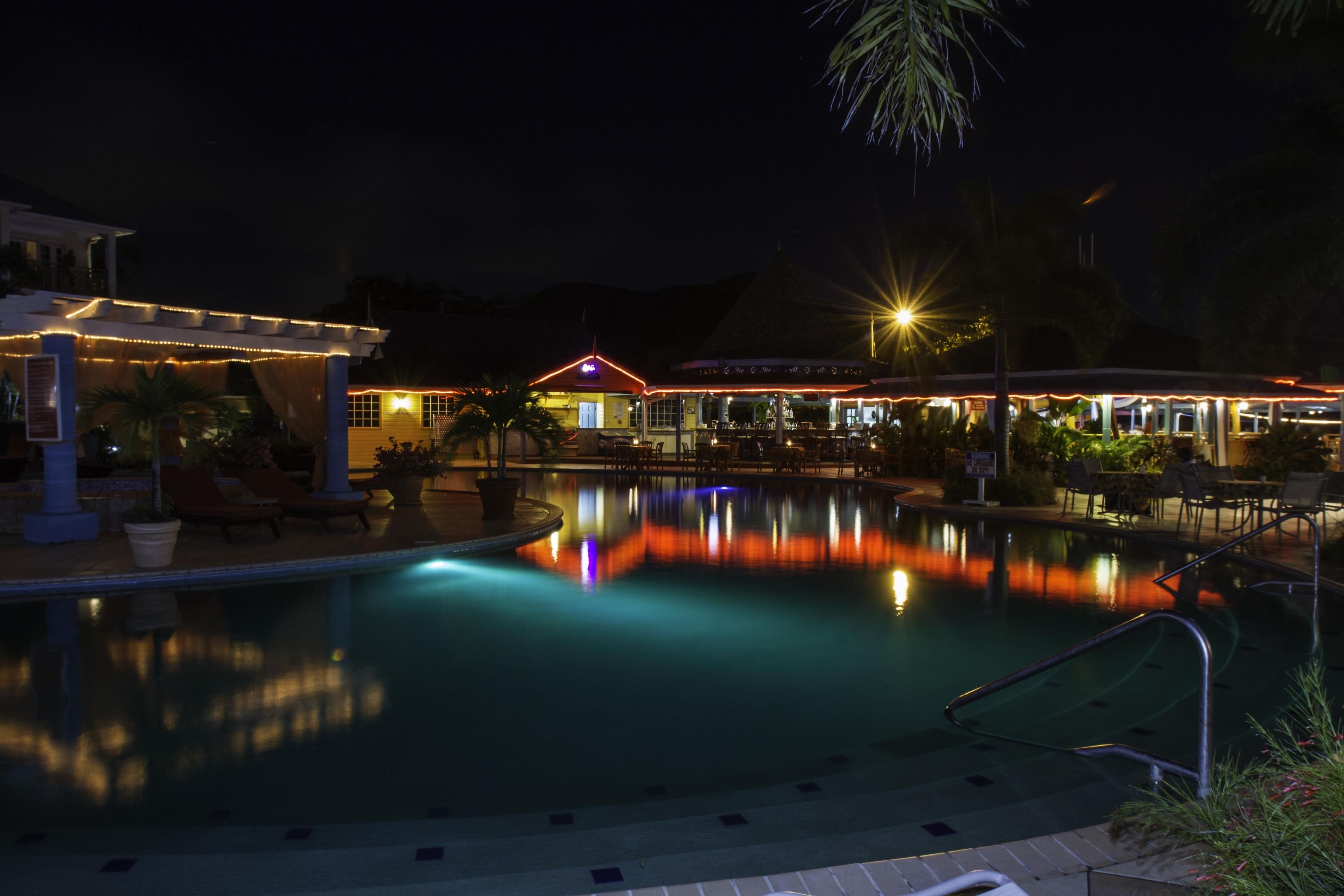 Un espace piscine illuminé la nuit, avec des jeux de lumière reflétés dans l’eau, des chaises longues et un bâtiment surmonté d’un toit rouge.