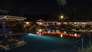 Un espace piscine illuminé la nuit, avec des jeux de lumière reflétés dans l’eau, des chaises longues et un bâtiment surmonté d’un toit rouge.