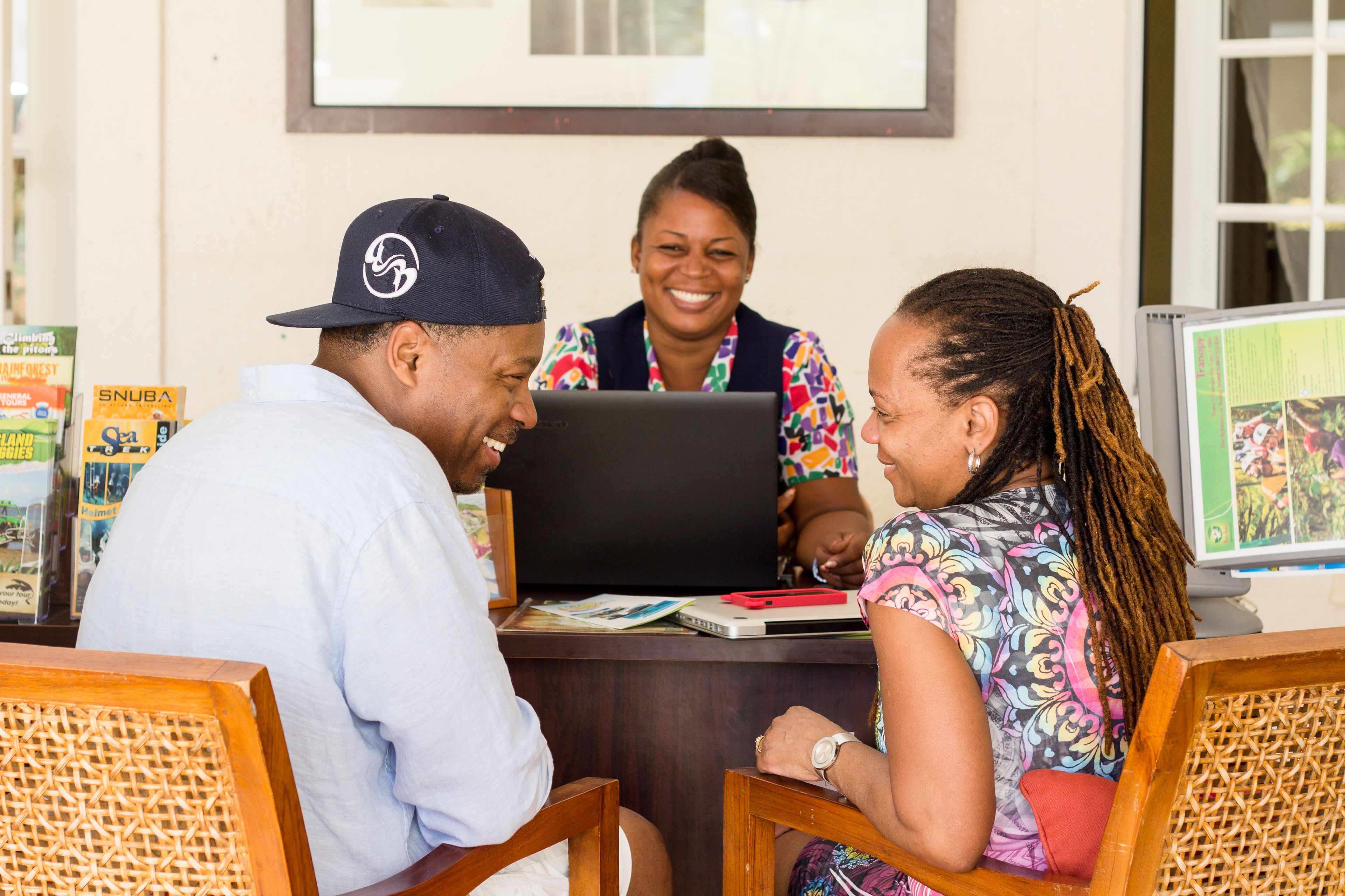 Three people sitting at a wooden table with a laptop, surrounded by magazines and a computer monitor.