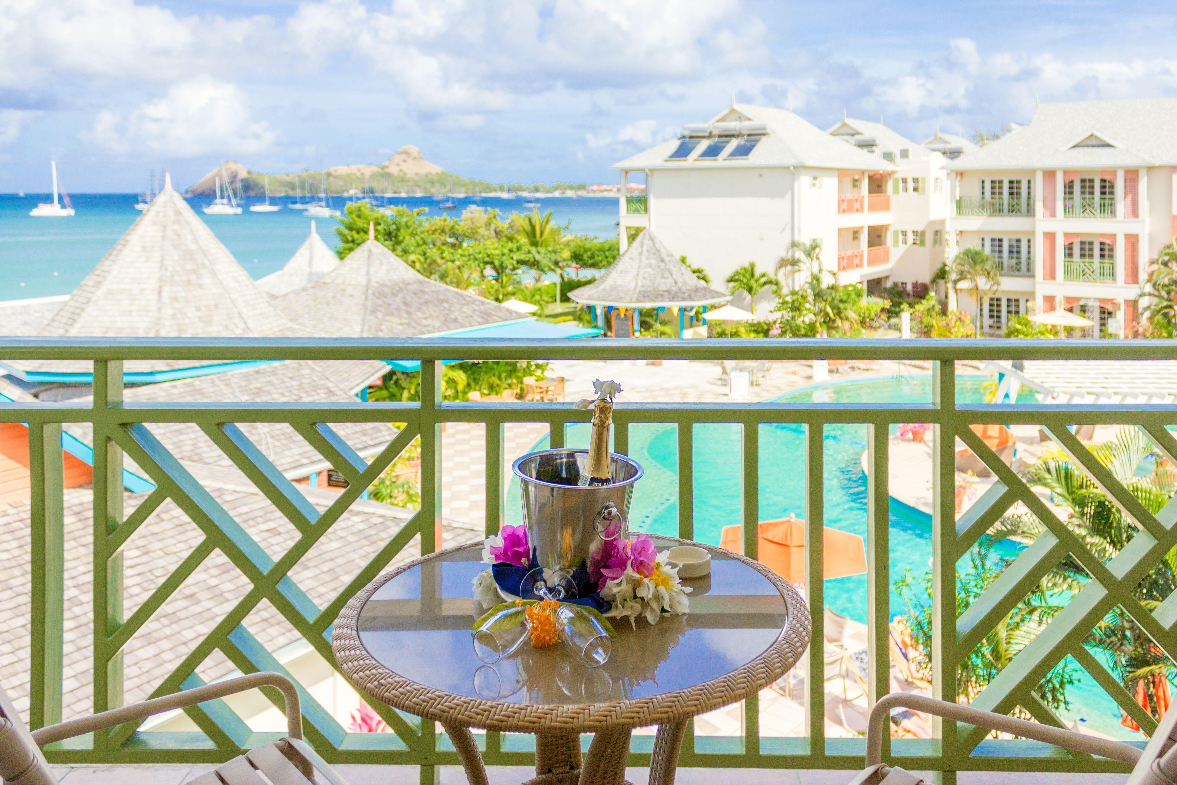 A balcony with a view of a pool, lounge chairs, and a tropical setting with buildings and a beach in the background.