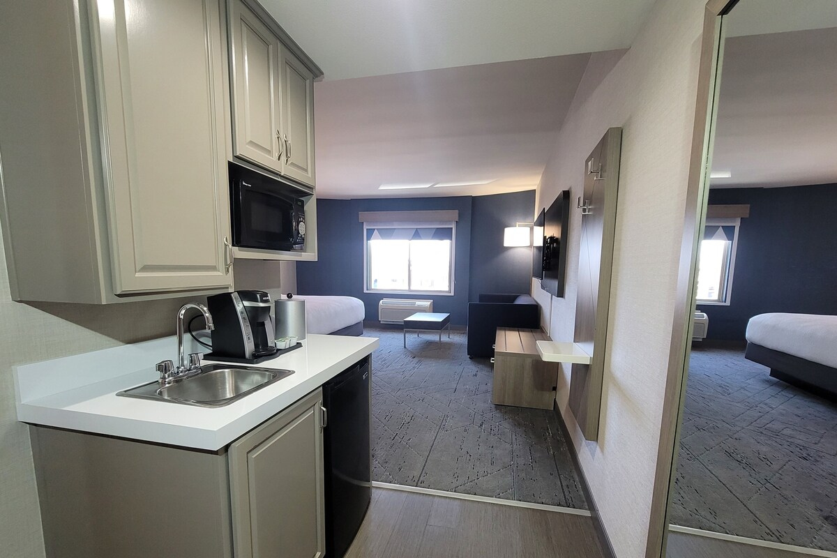 Habitación de hotel compacta con kitchenette, una cama, un pequeño rincón de descanso y una ventana que proporciona luz natural.