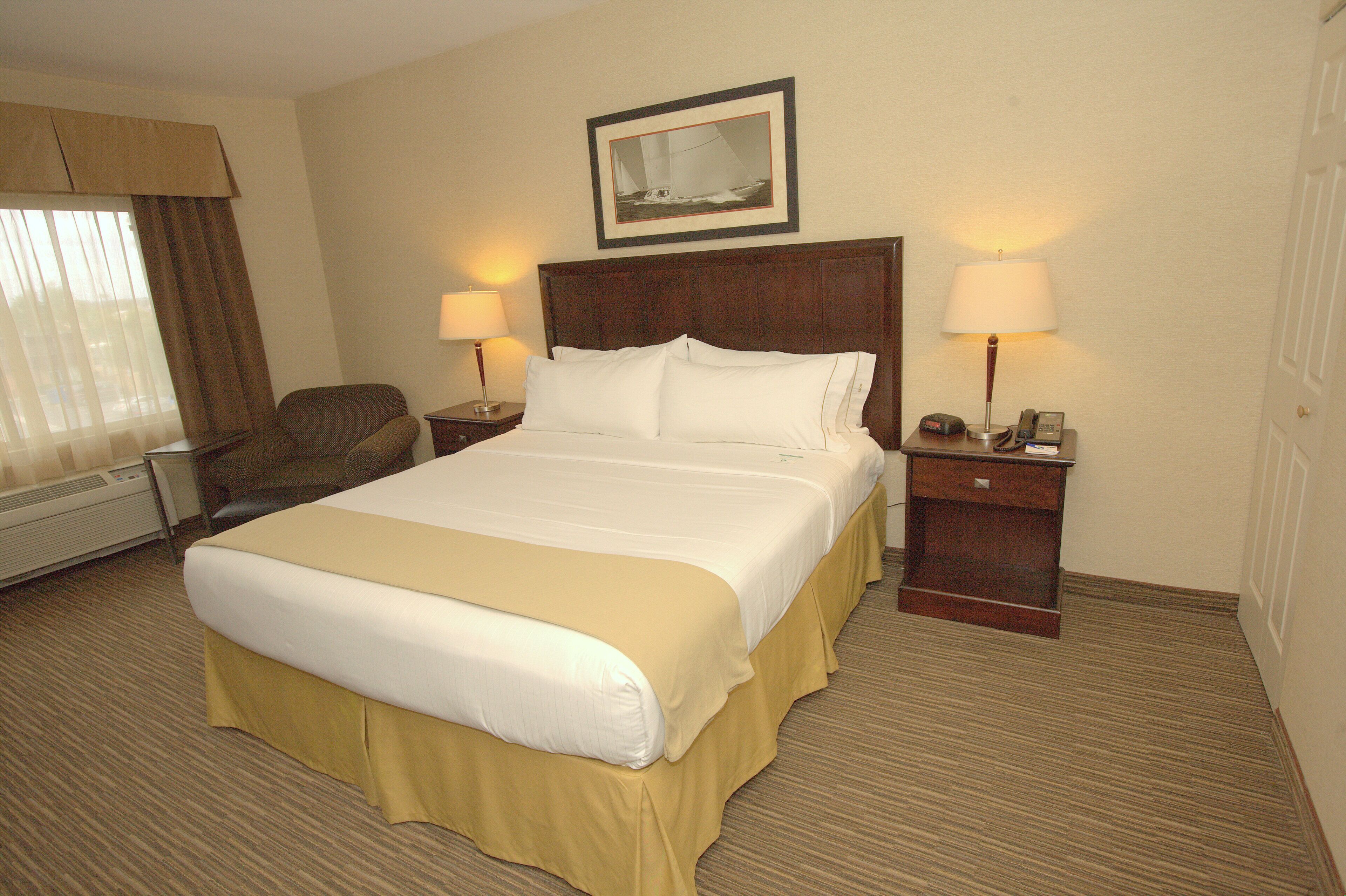 Quarto de hotel com cama grande, duas mesas de cabeceira com abajures, uma poltrona, janela com cortinas e um quadro na parede.
