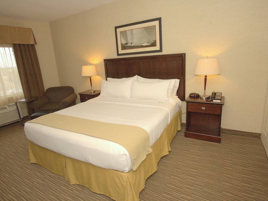 Quarto de hotel com cama grande, duas mesas de cabeceira com abajures, uma poltrona, janela com cortinas e um quadro na parede.