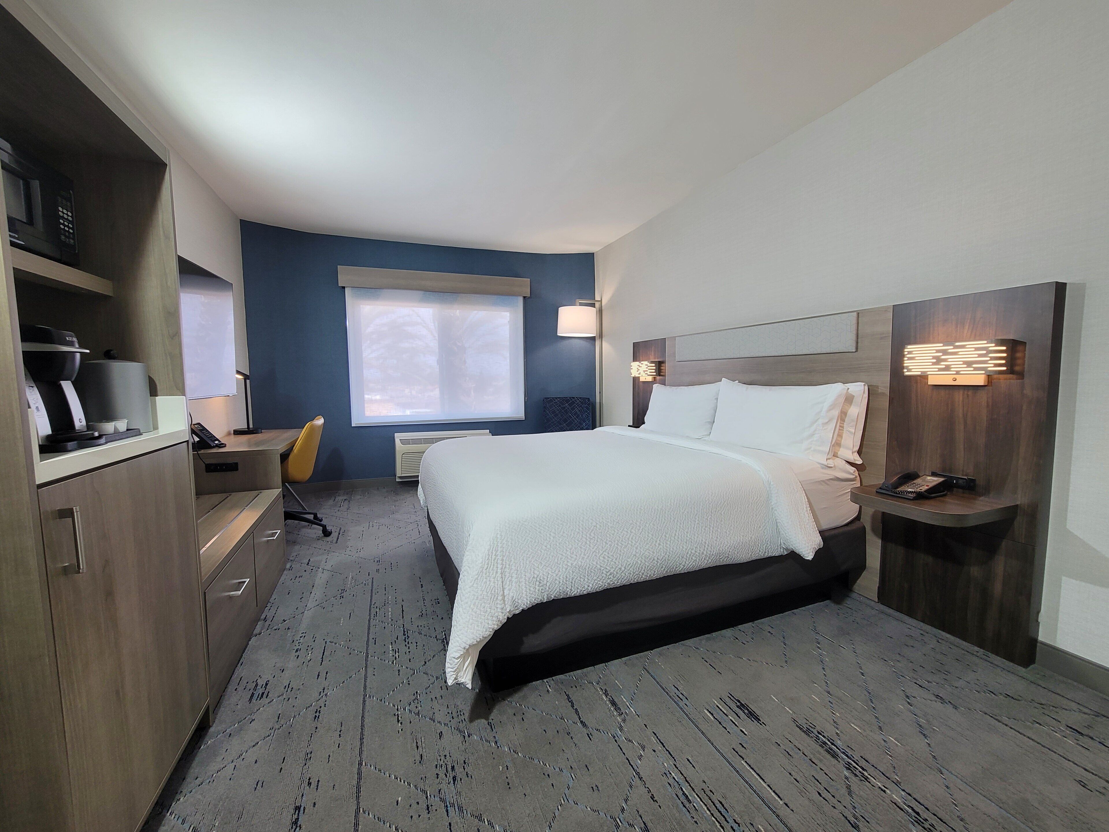 A modern hotel room with a large bed, a desk, a chair, and a window with blinds.