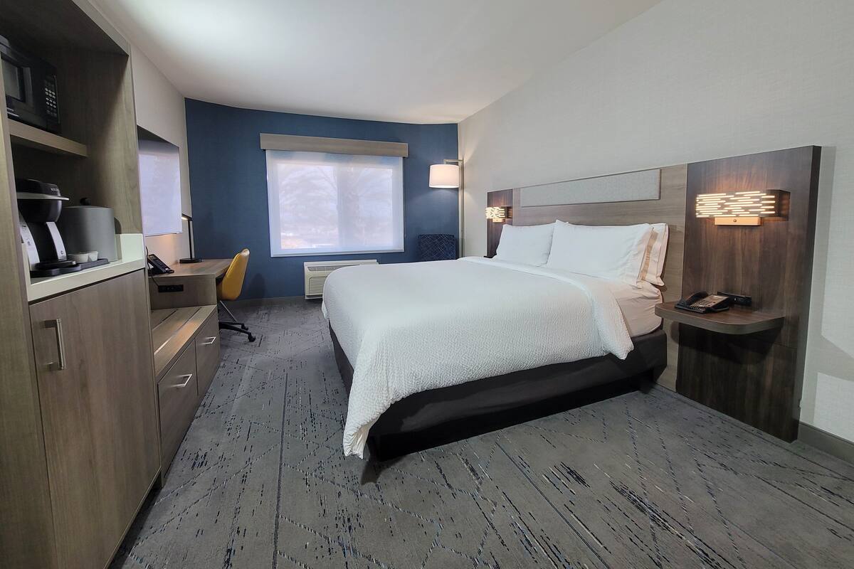 A modern hotel room with a large bed, a desk, a chair, and a window with blinds.