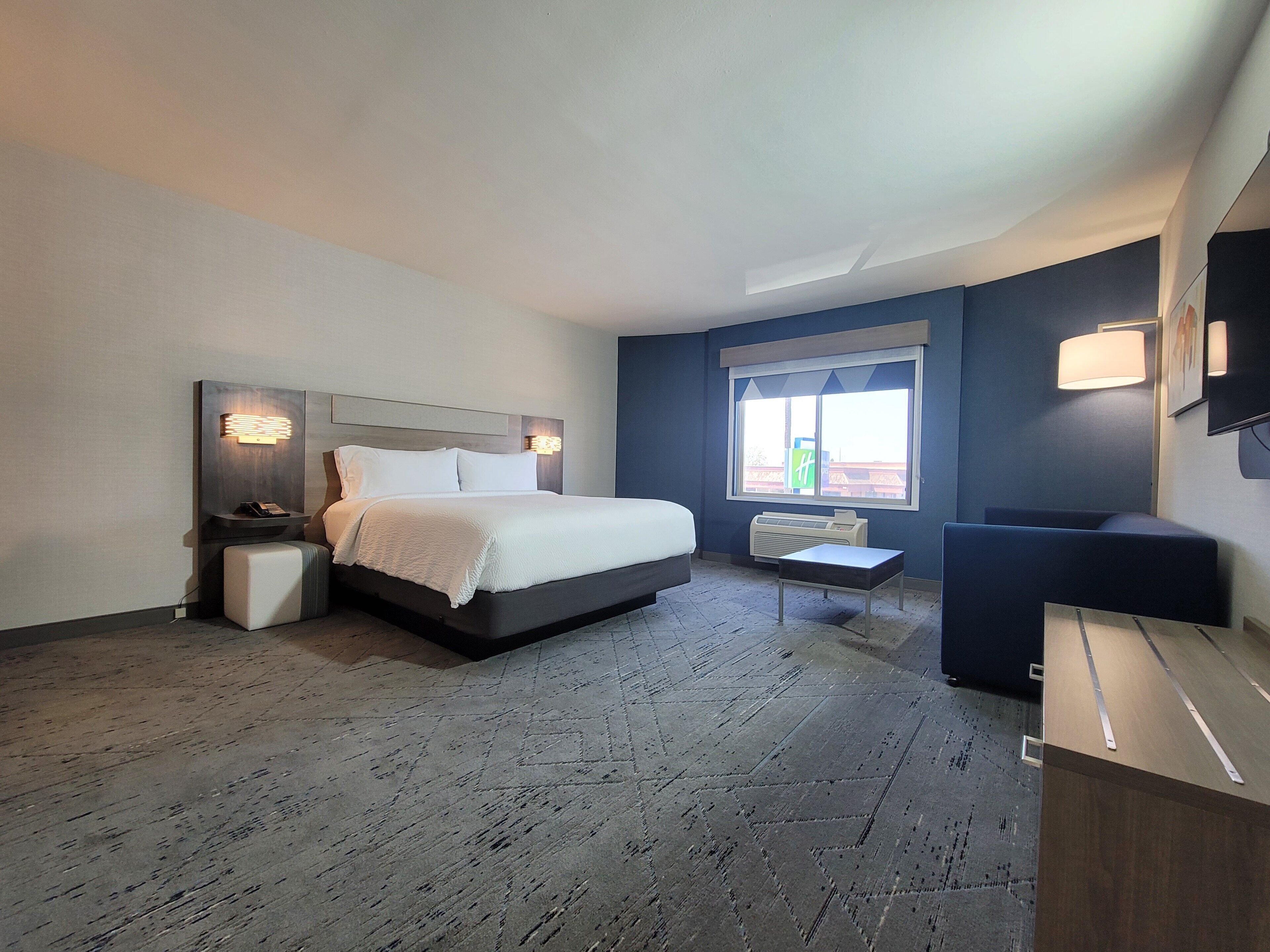 A modern hotel room with a large bed, bedside tables, a desk, a chair, a window, and a TV on the wall.