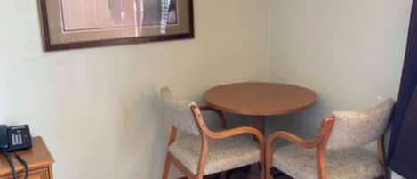 A small room with a round wooden table and two upholstered chairs. A framed picture hangs on the wall, and a telephone is on a wooden side table.