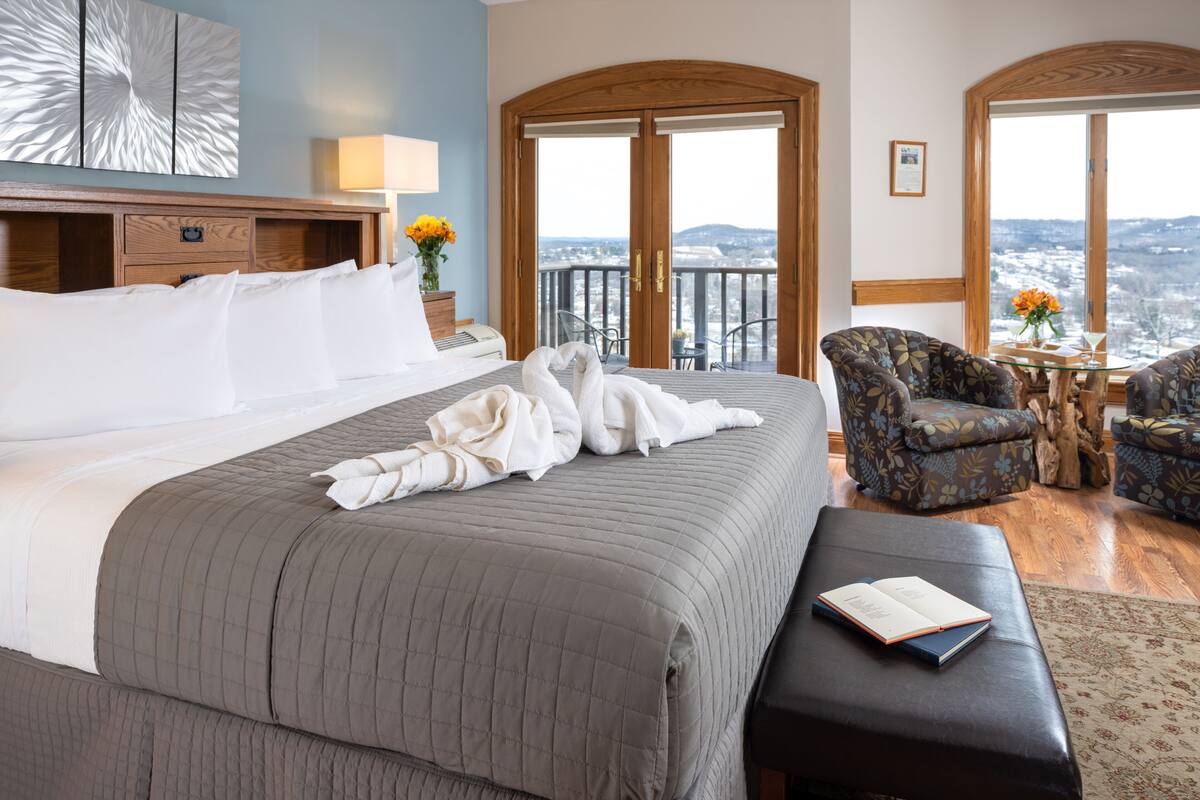 Una habitación de hotel con una cama grande, cabecera de madera, balcón con vistas y zona de estar con mesa y sillas.