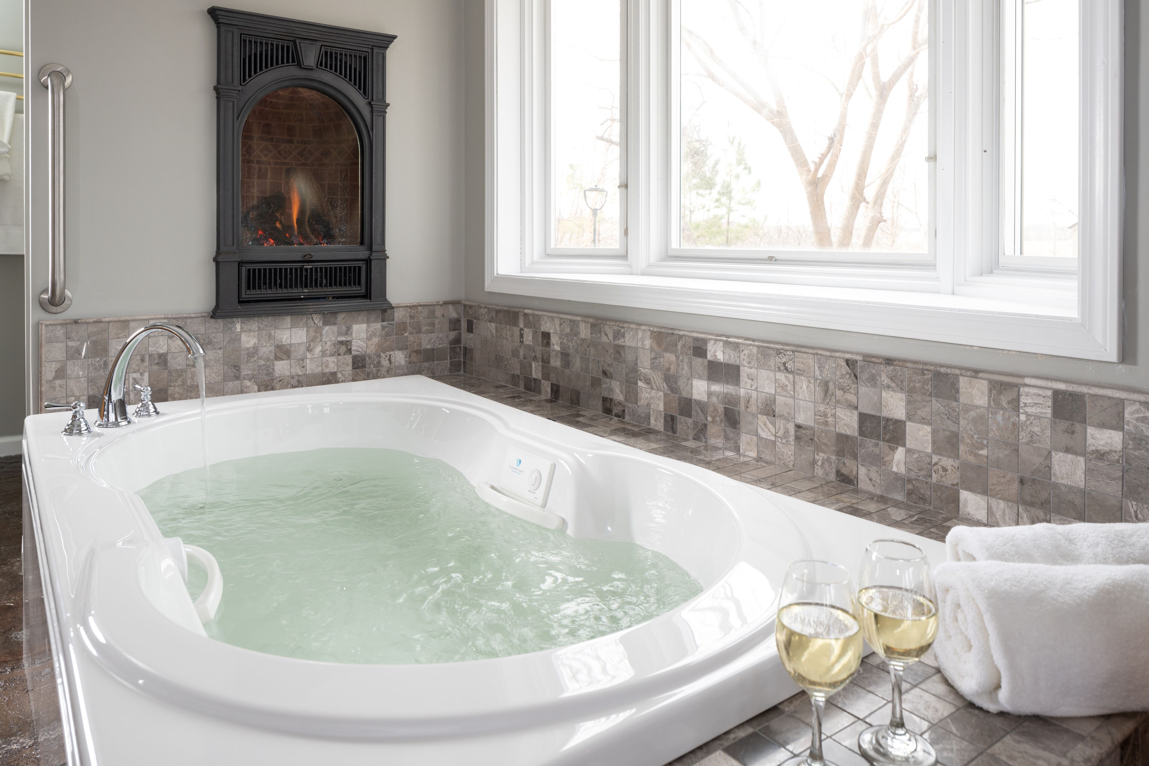 A bathroom with a large bathtub, a fireplace, and a window with a view of trees.