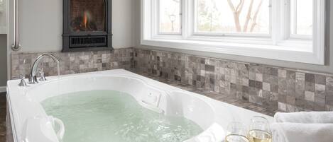 A bathroom with a large bathtub, a fireplace, and a window with a view of trees.