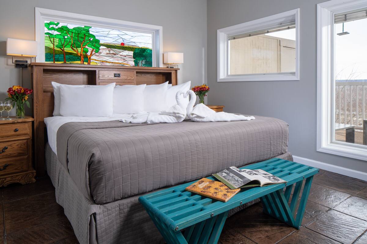 Una cama bien hecha con una colcha gris acolchada, una mesita de noche con una lámpara, una pequeña mesa con un florero y una ventana con vistas al exterior.