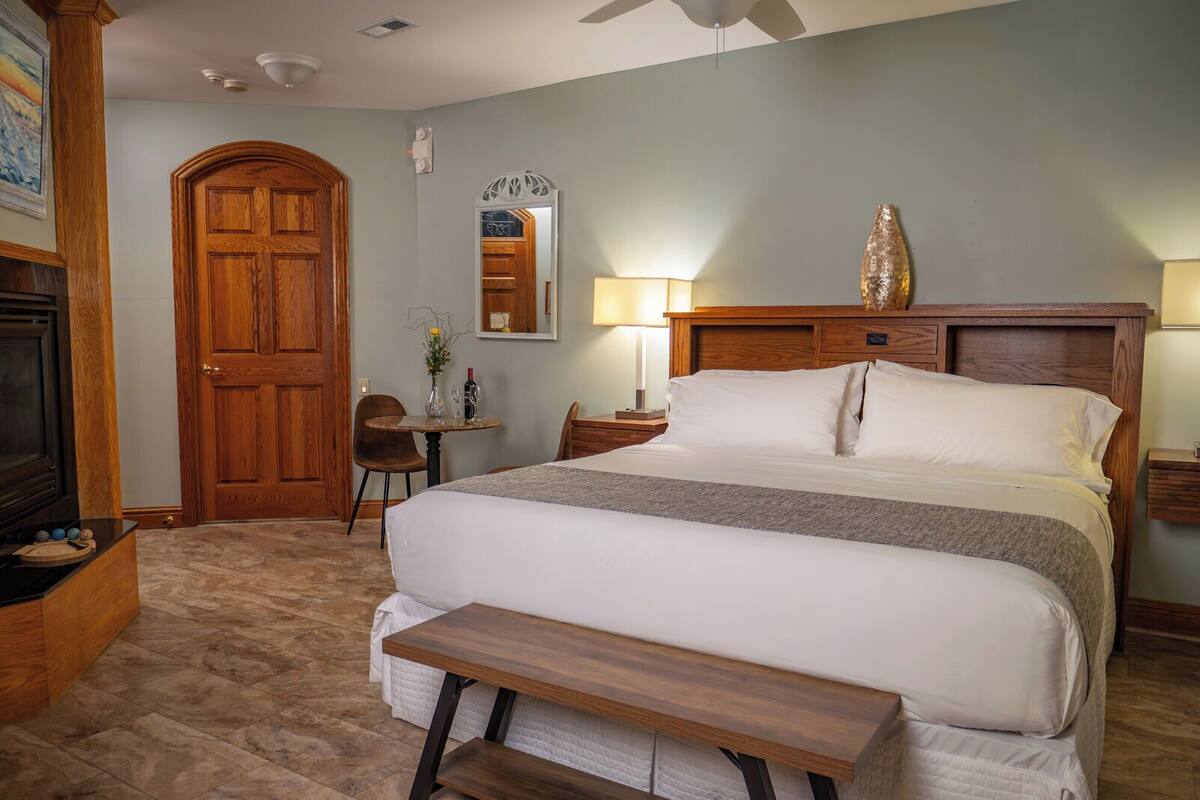 Habitación de hotel con una cama grande, cabecero de madera, mesitas de noche con lámparas, una pequeña mesa redonda con un florero y una botella de vino, un banco de madera y una puerta con un espejo encima.