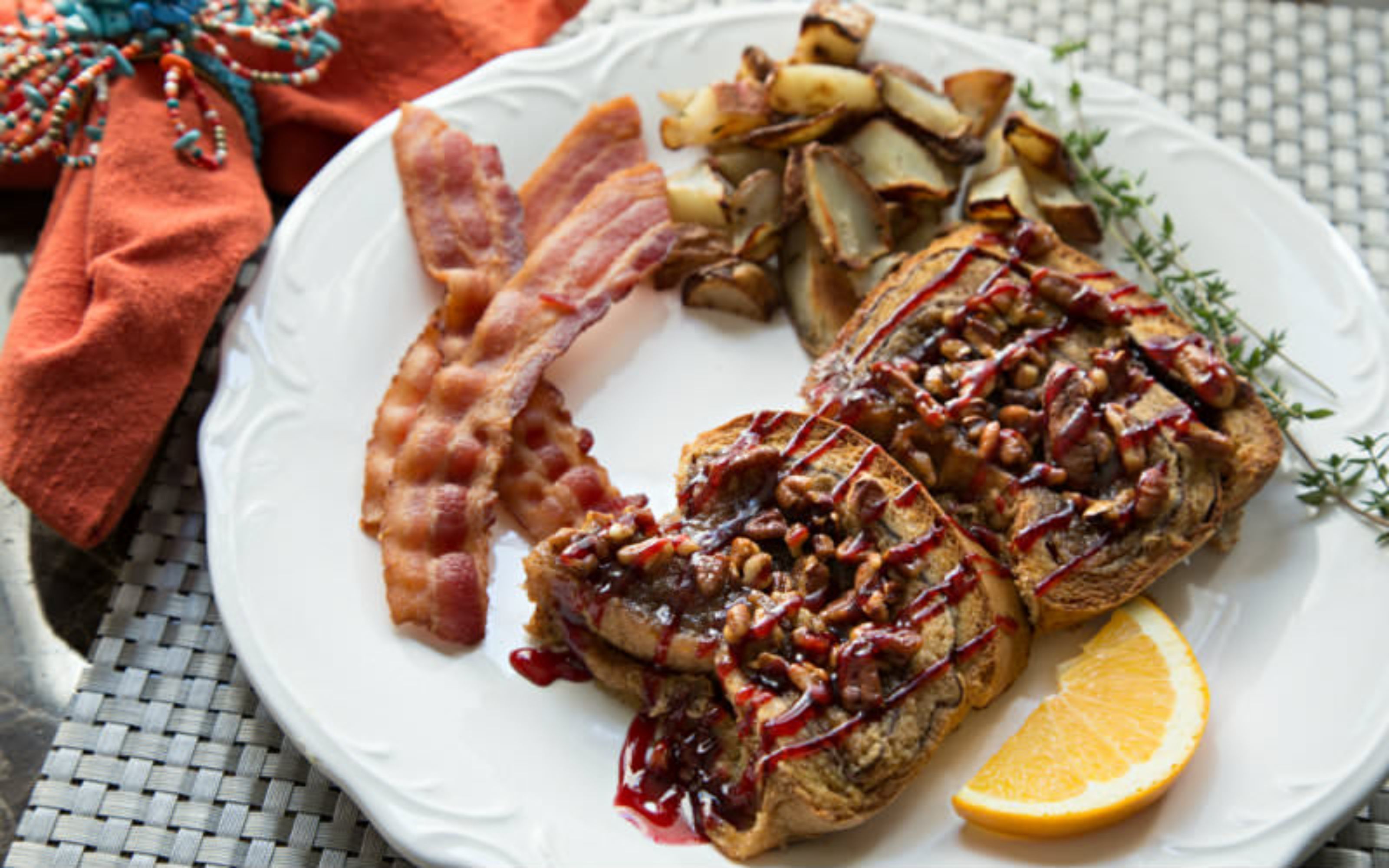 A plate with bacon, roasted potatoes, and a glazed meat dish, accompanied by an orange slice.