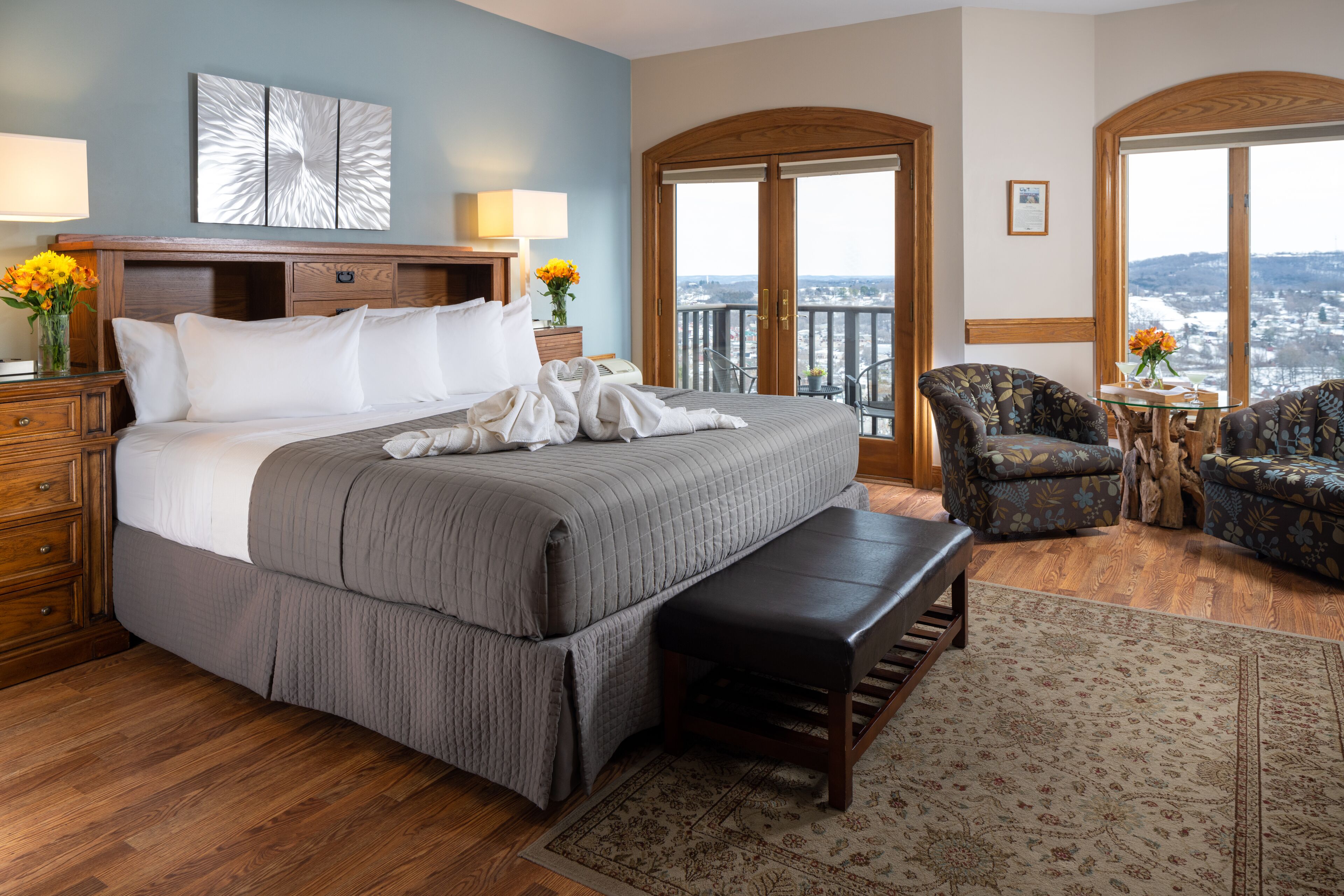 A hotel room with a large bed, a wooden bench, two armchairs, a view of the city through a balcony door, and a large window.