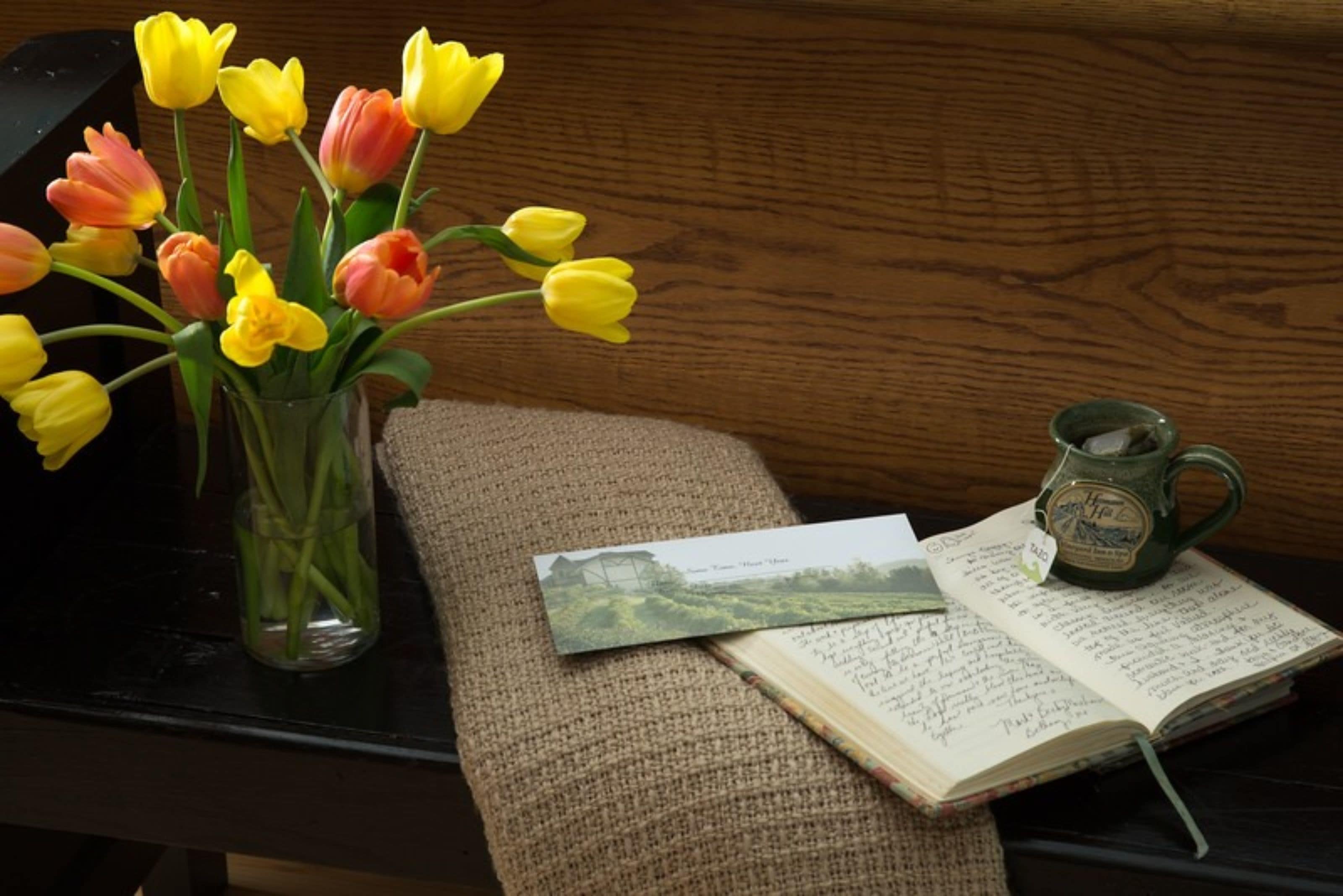 A wooden table with a vase of tulips, a mug, and a notebook with a picture.