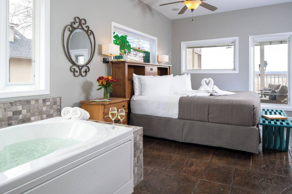 Habitación de hotel con una bañera grande, una cama con colcha gris, una mesita de noche de madera con un florero y un cuadro de un árbol.