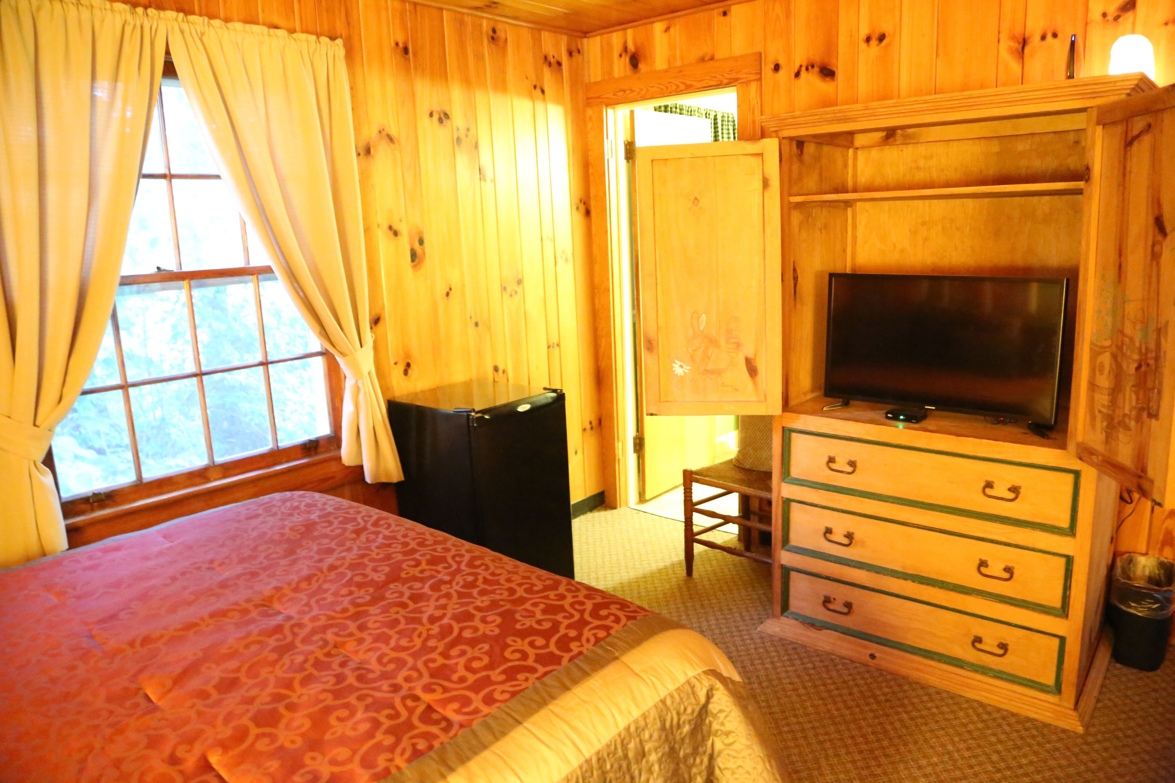A wooden room with a bed, a TV on a dresser, and a window with curtains.