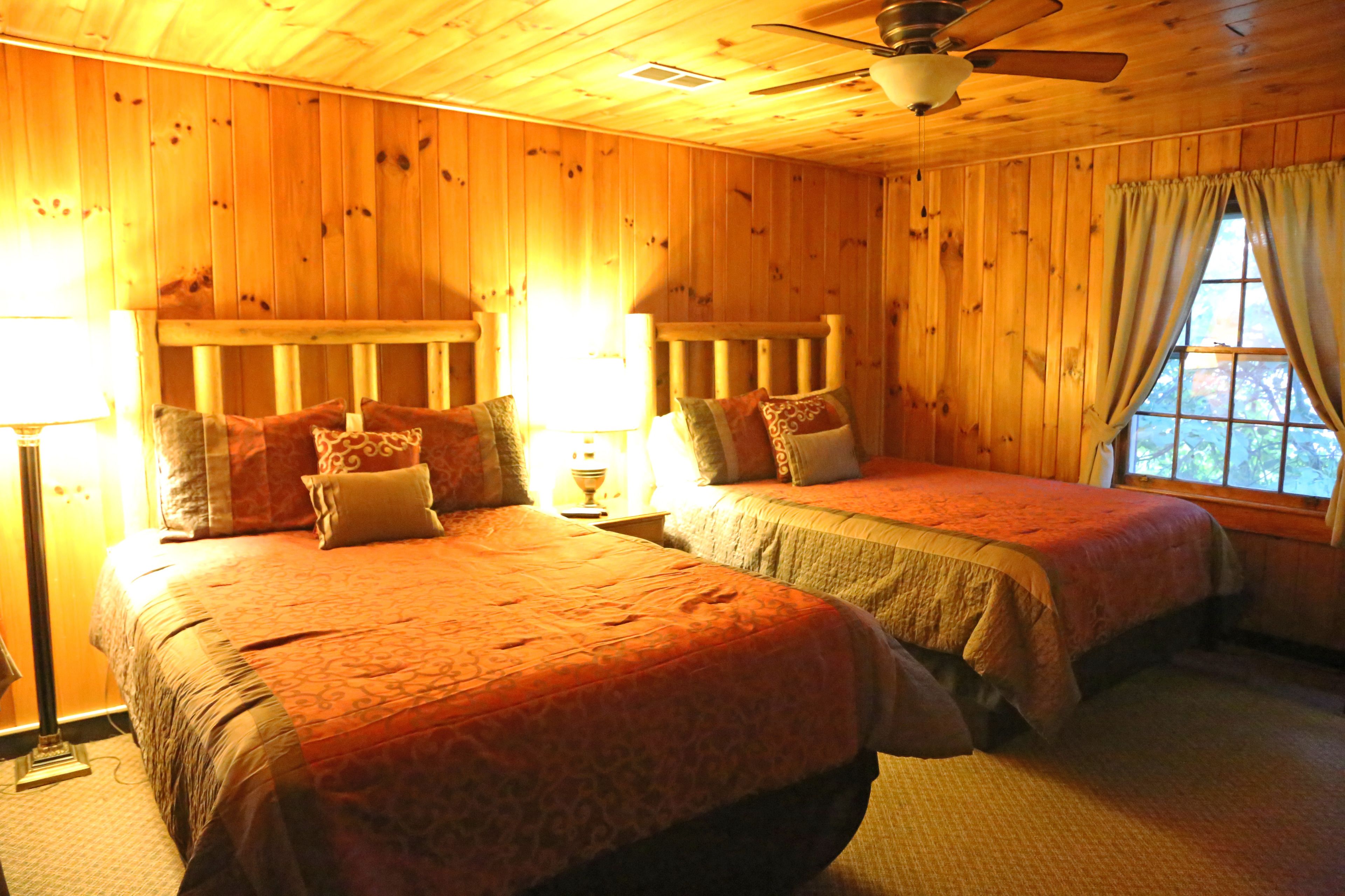 A room with two beds, wooden walls, a ceiling fan, and a window with curtains.