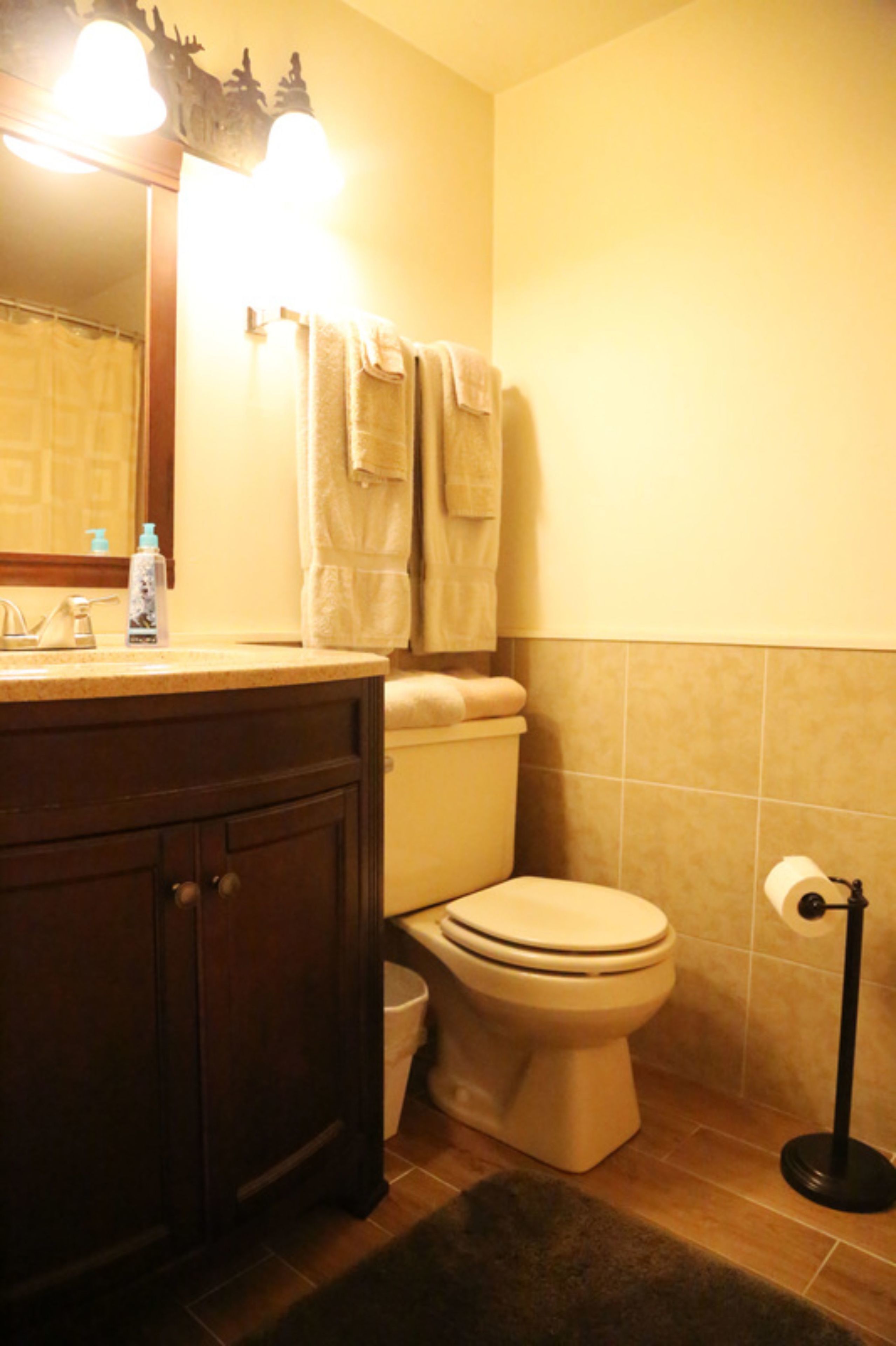 Ein Badezimmer mit Toilette, Waschbecken und Spiegel. Handtücher hängen an der Wand und ein Mülleimer steht bereit.