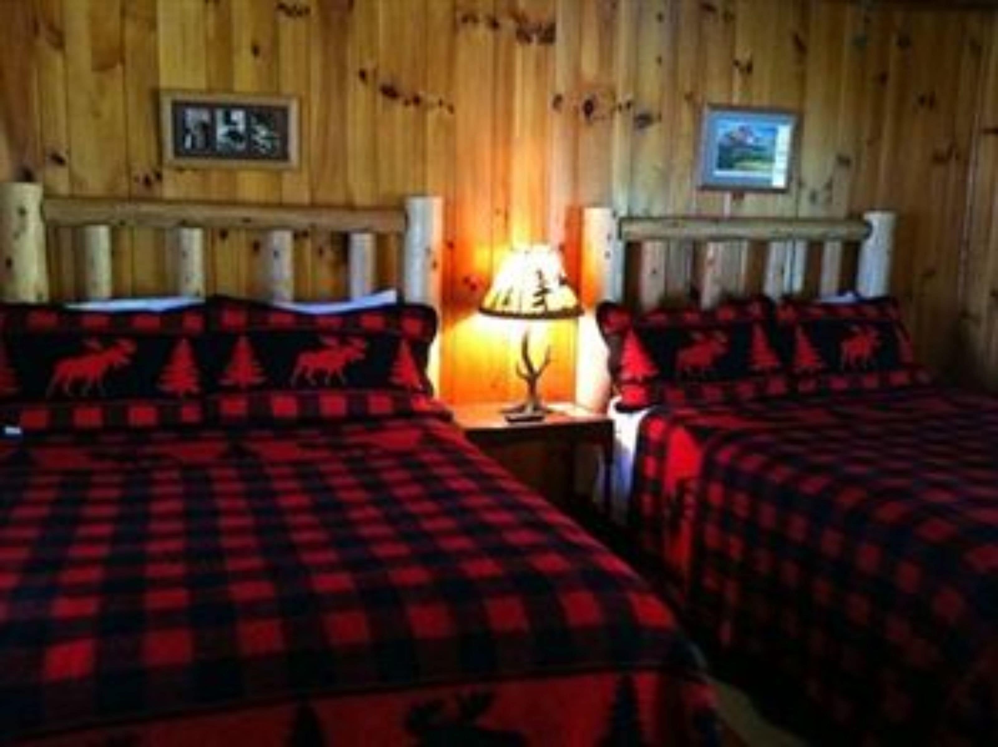 A room with two beds, each with red and black checkered bedding and a red animal motif. There is a nightstand with a lamp between the beds.