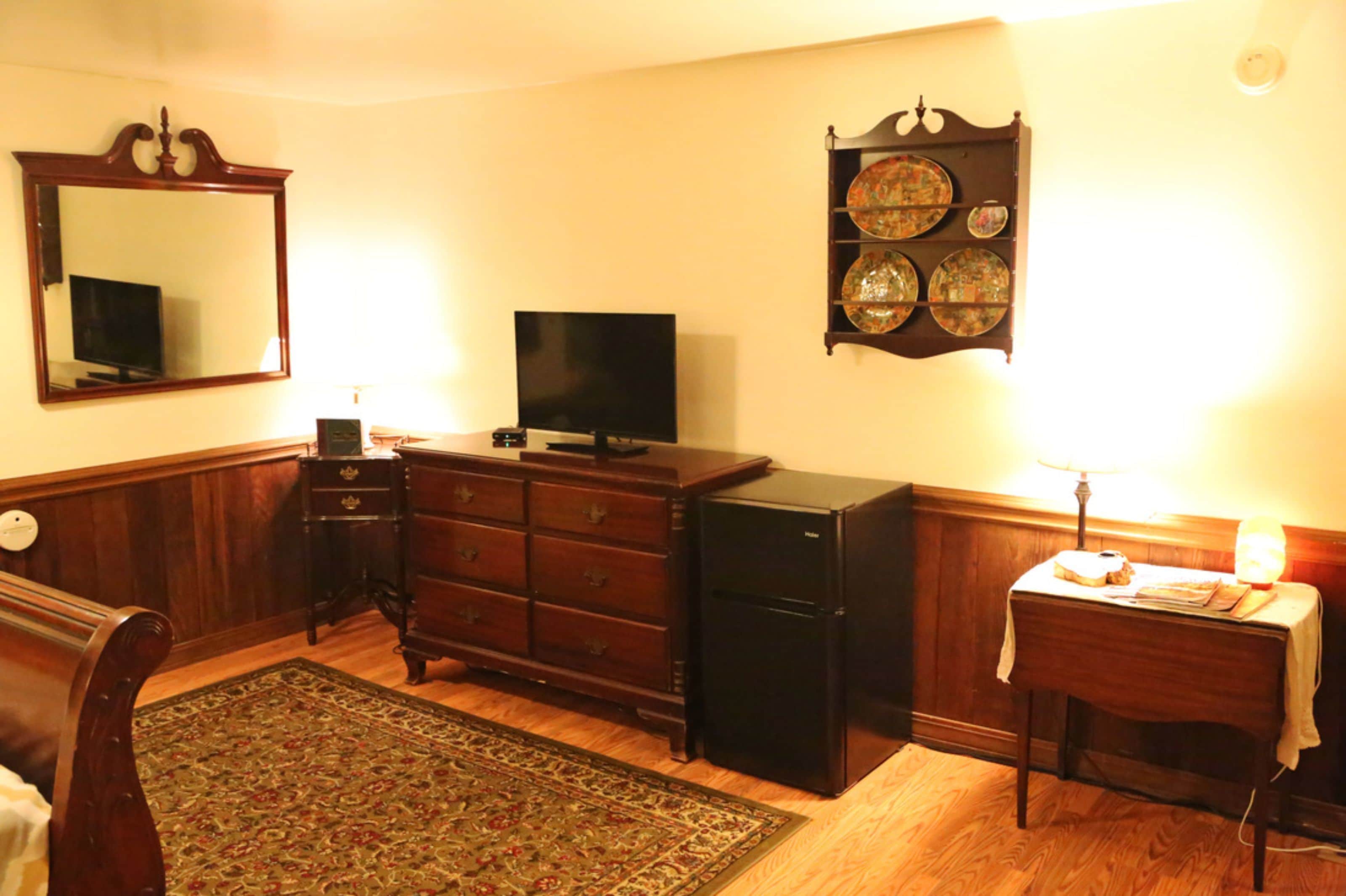 Une chambre comprenant une commode en bois, un téléviseur, un petit réfrigérateur, un miroir et un porte-assiettes fixé au mur.