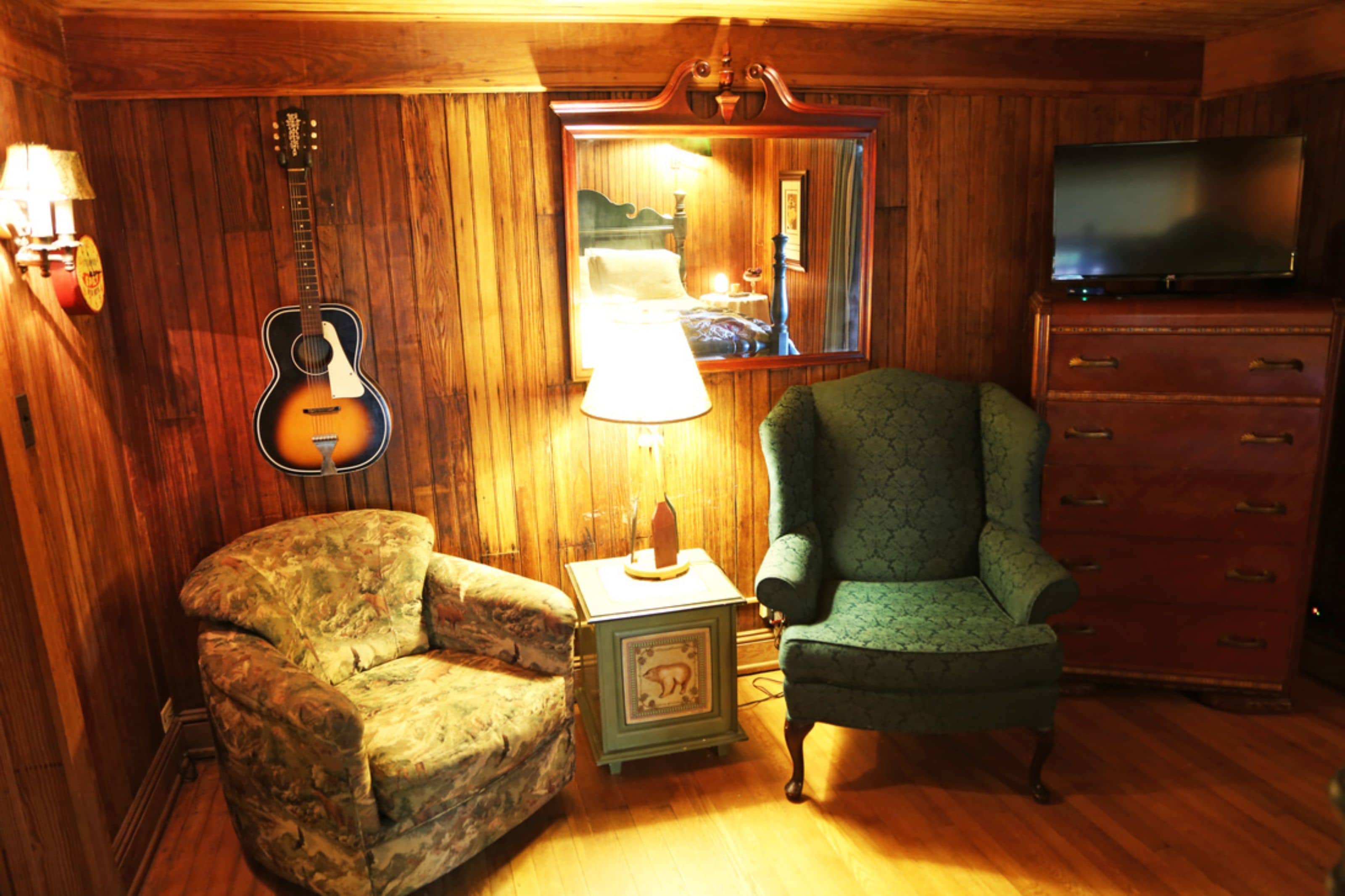 A room with wooden walls, a guitar, a lamp, a chair, a dresser, and a television.