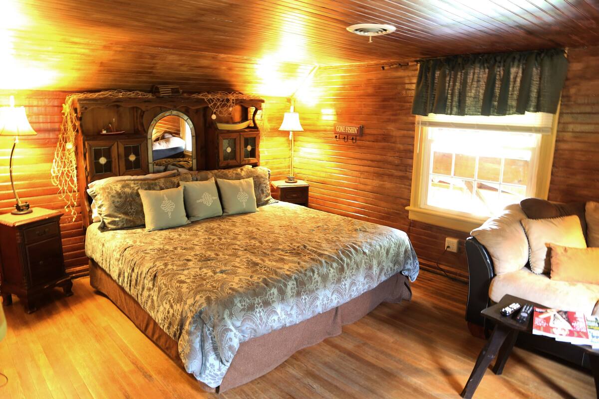 A cozy wooden cabin bedroom with a large bed, two bedside tables, a chair, and a window with curtains.