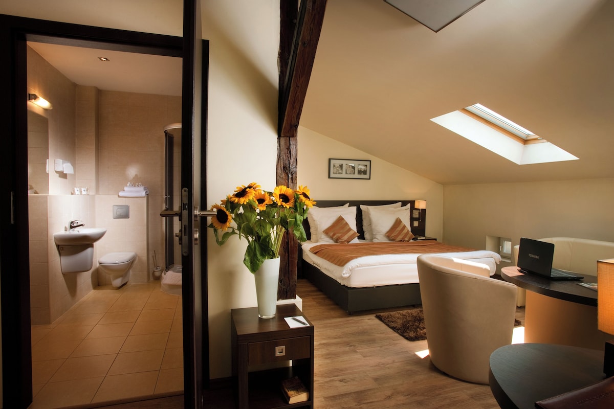Une chambre avec une lucarne, une table de chevet sur laquelle se trouve un ordinateur portable, une chaise et une petite table de nuit ornée d’un vase de tournesols.