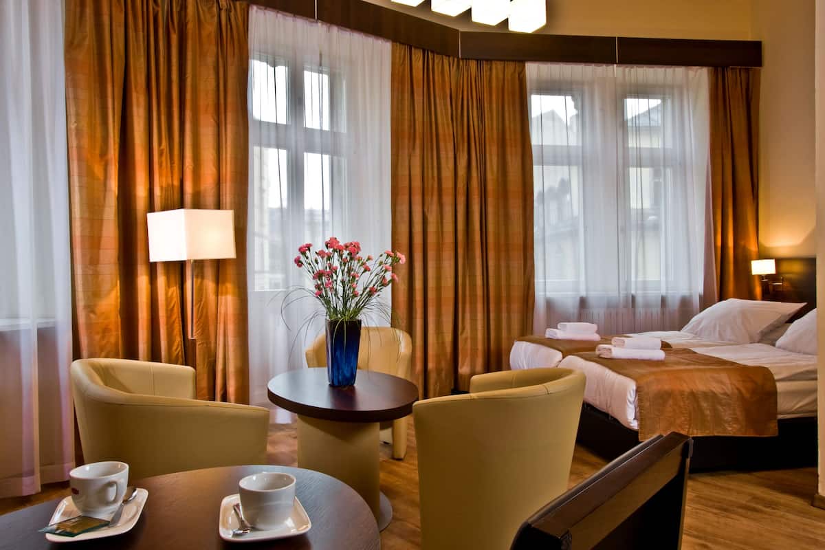 A hotel room with a bed, two chairs, a table with cups and saucers, a vase with flowers, and curtains.