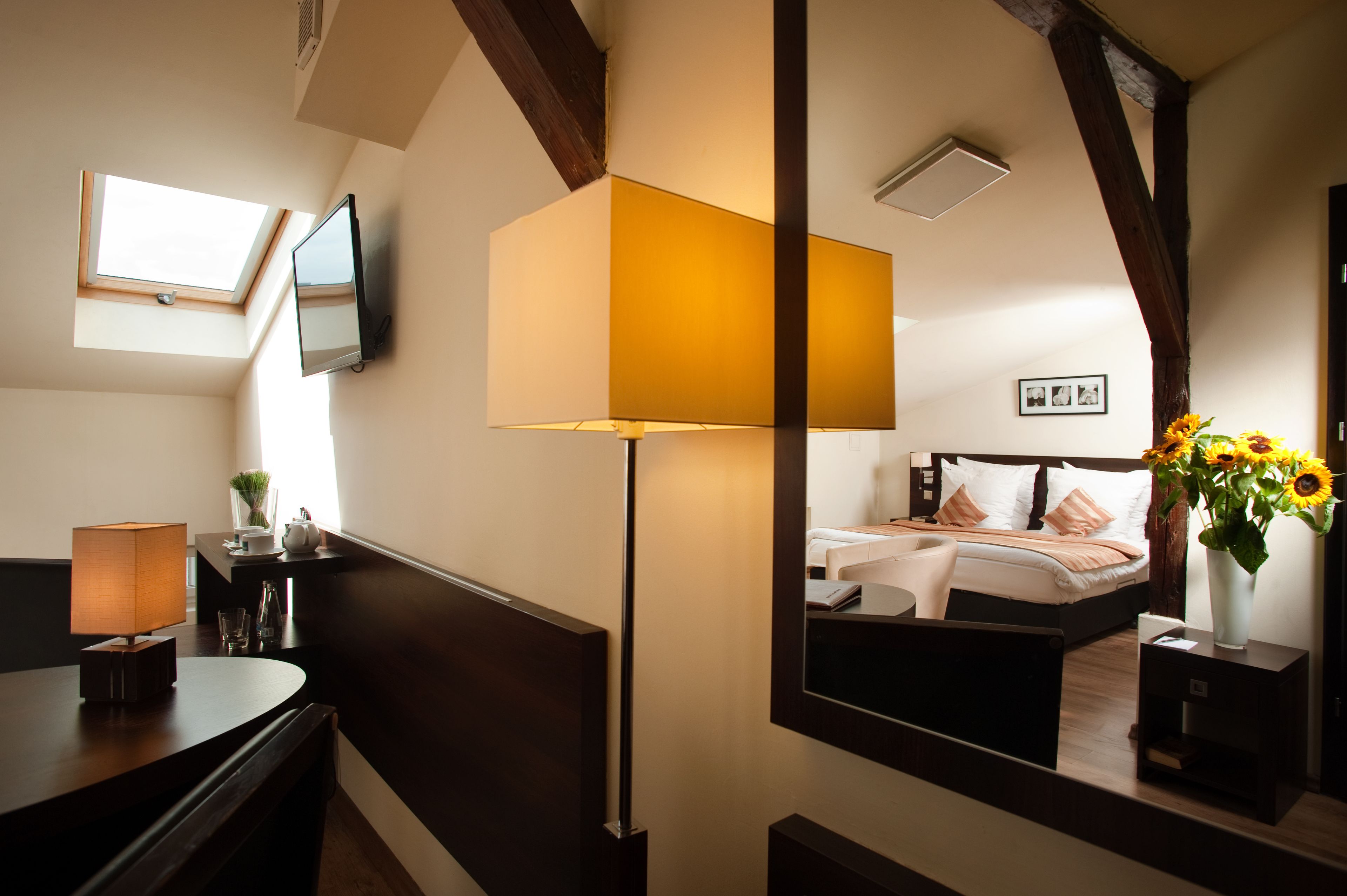 A hotel room with a bed, bedside table, lamp, and a large mirror reflecting the room.