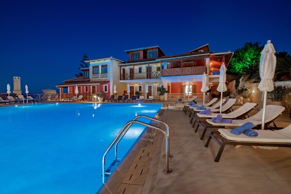 A swimming pool with lounge chairs and umbrellas, a multi-story building with balconies, and a stone wall. - Sea View Village - Studios & Apartments (Zakynthos)