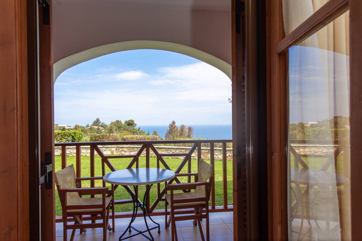 An open balcony with a table and chairs, offering a view of the sea and greenery.