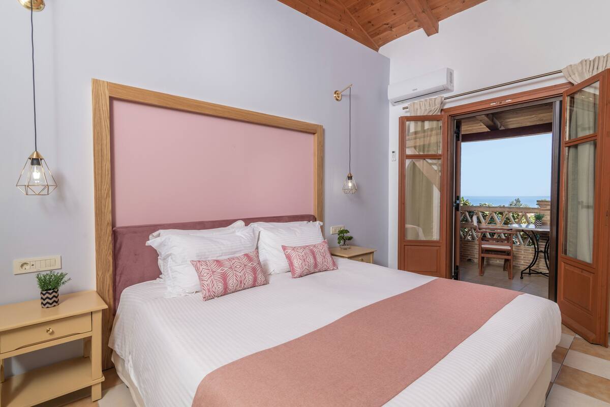 A bedroom with a large bed, a wooden headboard, a nightstand, a wall-mounted lamp, and a view of an outdoor patio.