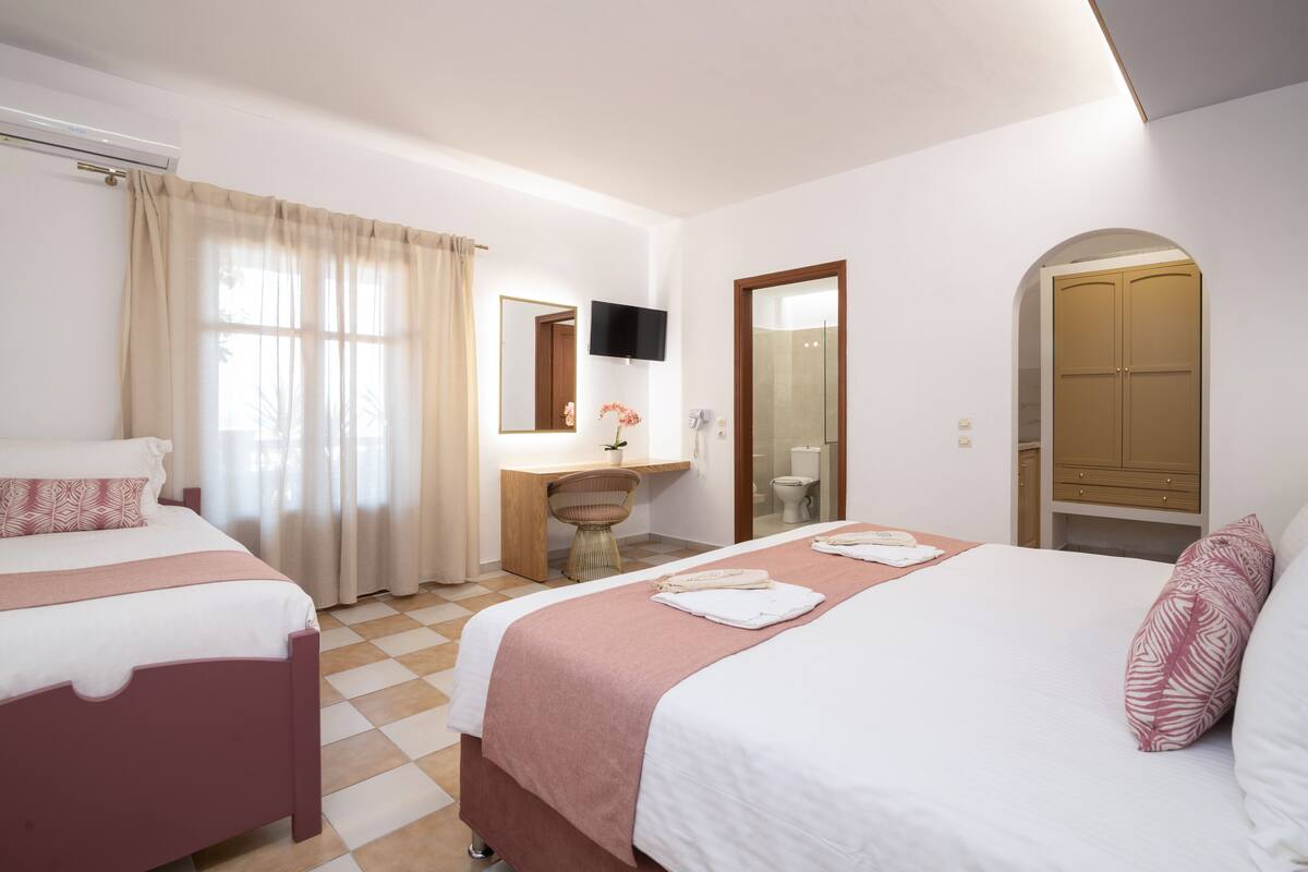 A hotel room with two beds, a desk, a TV, and a bathroom visible through an archway.