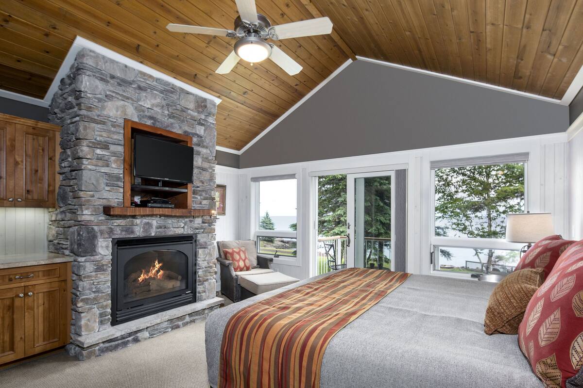 A bedroom with a stone fireplace, a bed, a television, and a balcony with a view.