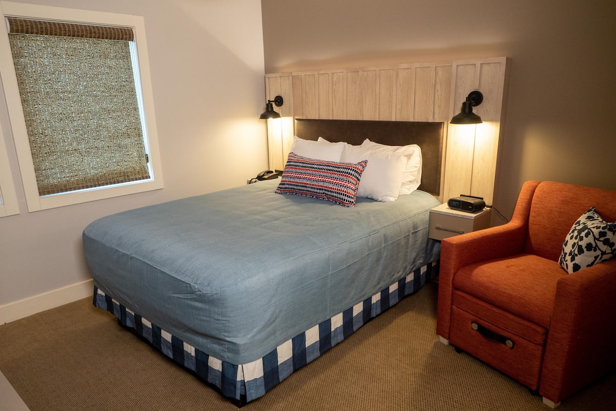 A neatly made bed with a blue bedspread and a checkered skirt, a wooden headboard, two bedside lamps, a telephone, and a red armchair with a patterned pillow.