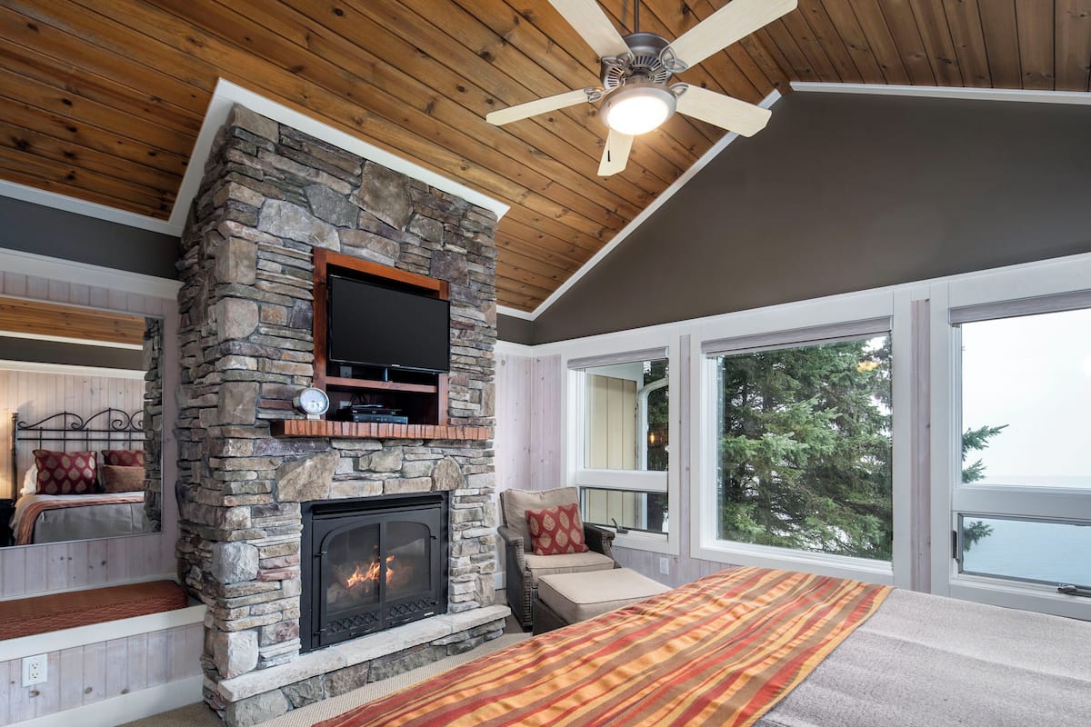A cozy living room with a stone fireplace, a flat-screen TV, and a large window offering a view of trees.