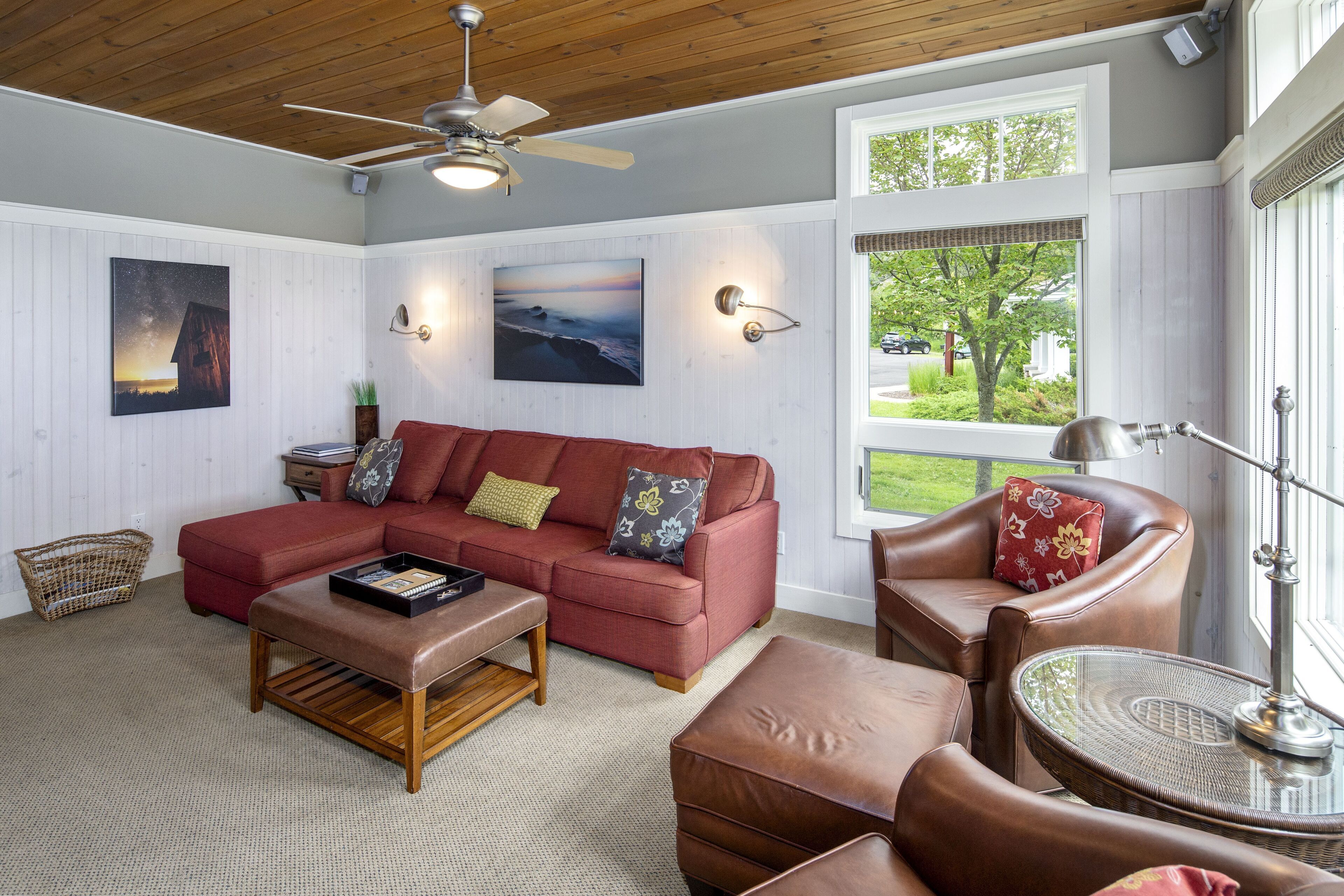 Una sala de estar acogedora con un sofá sectional rojo, un sillón de cuero marrón, una mesa de centro de madera y una mesita auxiliar de mimbre.