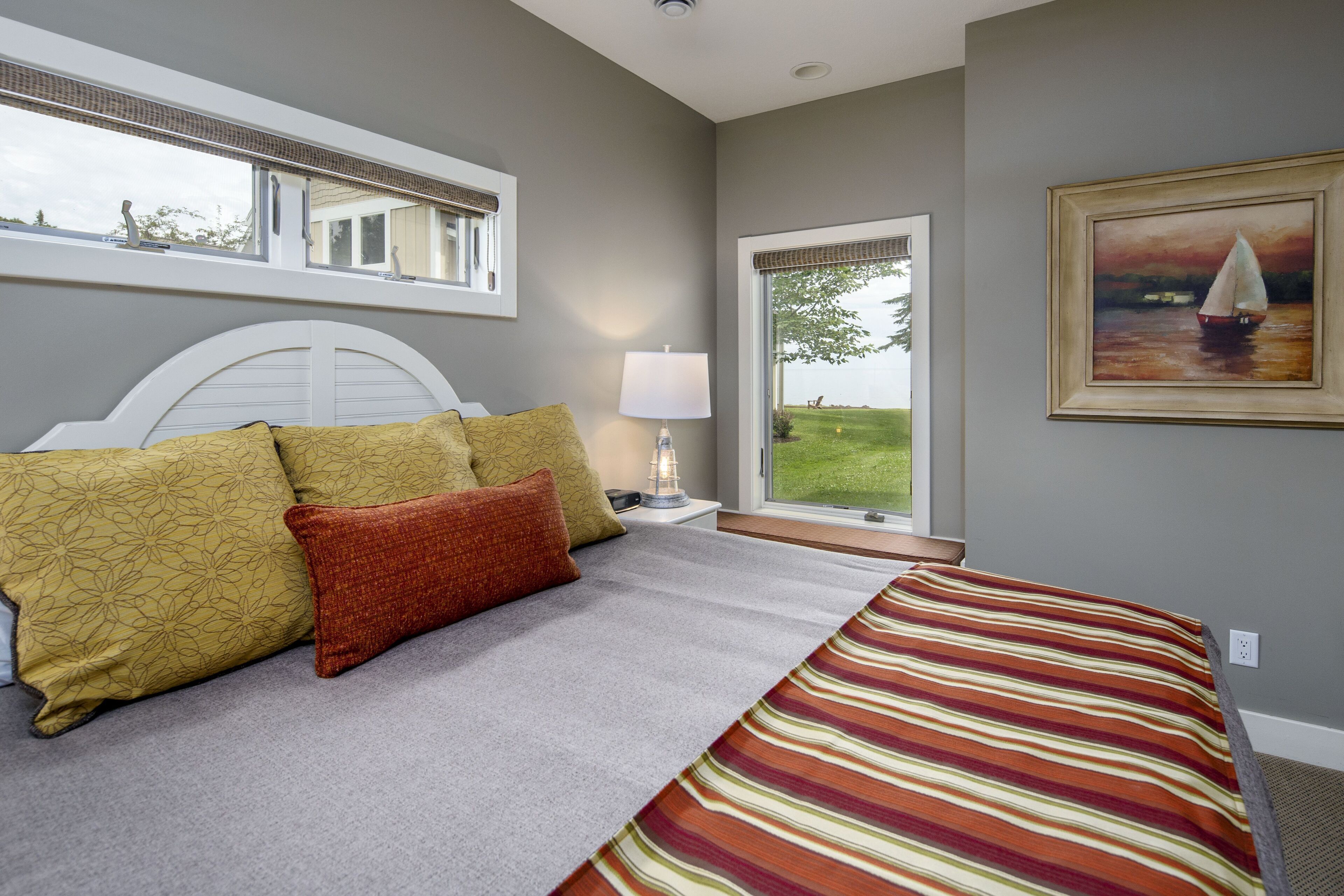 A bedroom with a bed, pillows, a nightstand, a lamp, a window with a view, and a painting on the wall.
