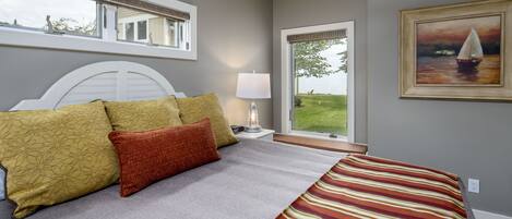 A bedroom with a bed, pillows, a nightstand, a lamp, a window with a view, and a painting on the wall.